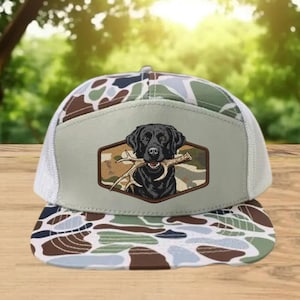 May include: A camouflage trucker hat with a black Labrador Retriever graphic holding antlers. The hat features a white mesh back, a patterned brim, and a tan front panel with the dog illustration. The hat is designed for outdoor enthusiasts.