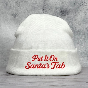 May include: A white knit beanie with a folded brim. The text "Put It On Santa's Tab" is embroidered in red on the brim. The hat is made of a soft, textured material, perfect for winter wear.