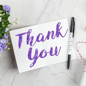 Może przedstawiać: Biała karta z fioletowym napisem "Thank You". Obok karty leży czarno-biały długopis z gwiazdkami. Obecny jest r&oacute;wnież mały prezent owinięty w biały papier z czerwoną i białą sznurkiem.