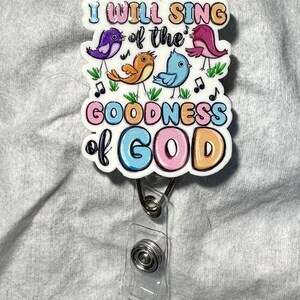 Op de afbeelding: Een kleurrijke badge reel met de tekst "I WILL SING of the GOODNESS of GOD". Het ontwerp toont vier cartoonvogels, muzieknoten en een bloemenrand. De reel heeft een doorzichtige plastic band en een metalen clip.
