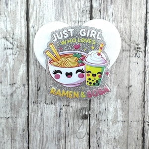 Puede incluir: Un portatarjetas con un borde plateado brillante que presenta un cuenco de ramen de dibujos animados y una bebida boba. El texto dice "Just Girl Who Loves Ramen & Boba". El carrete tiene un respaldo en forma de corazón blanco y un cordón retráctil con un clip de metal.