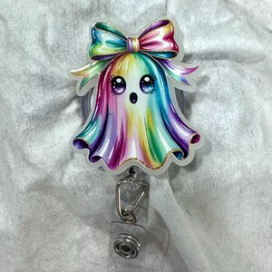 May include: A colorful badge reel featuring a cartoon ghost with a rainbow-colored body and a large bow. The ghost has large eyes and a surprised expression. The reel has a clear plastic strap and a metal clip.