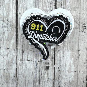 Könnte beinhalten: Ein herzförmiges Ausweis-Jojo mit den Worten "911 Dispatcher" in einer stilisierten Schriftart. Das Design enthält eine Kopfhörergrafik und die Nummer "911" in Gelb. Der Rand enthält Wörter wie "nett", "zuverlässig" und "mitfühlend".