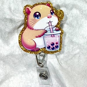 May include: A cute hamster badge reel with a gold glitter border. The hamster is drinking a pink bubble tea with dark purple boba pearls. The badge reel has a clear plastic strap and a metal clip.