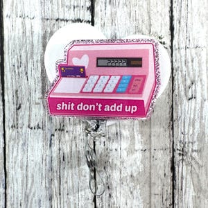 Peut inclure: Un porte-badge rose et scintillant avec un design de caisse enregistreuse et le texte "shit don't add up". Le porte-badge a un dos blanc et un clip argenté. La caisse enregistreuse comprend une fente pour carte de crédit et un cœur.