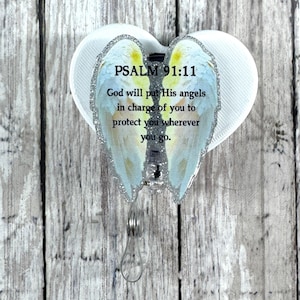 May include: A heart-shaped badge reel featuring a pair of light blue and yellow angel wings with silver glitter edges. The text reads: PSALM 91:11 God will put His angels in charge of you to protect you wherever you go. The reel has a clear plastic strap.