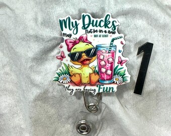 May include: A badge reel with a cartoon duck wearing sunglasses and a pink bow. The text reads "My Ducks may not be in a row, but at least they are having Fun." The reel also features a pink drink with ice cubes and butterflies.