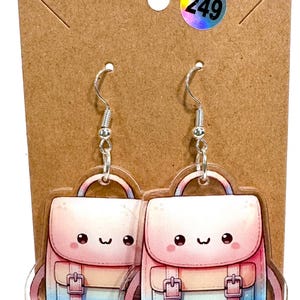 May include: A pair of silver-tone dangle earrings featuring a backpack design. The backpacks are in shades of pink, blue, and white, with a smiling face and silver-tone buckles. The earrings are attached to a brown cardboard backing.