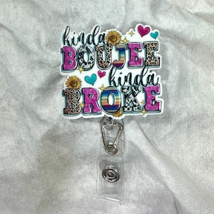 May include: A white badge reel with the text "kinda BOUJEE kinda BROKE" in a colorful design. The text features leopard print, floral, and southwestern patterns, with accents of hearts and stars. The reel has a clear plastic holder and a metal clip.