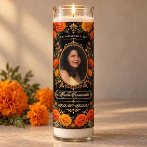 May include: A tall cylindrical glass prayer candle with a white wax core and a lit wick. The label features a black background with orange marigold flowers, a central oval portrait of a woman, and the text EN MEMORIA DE Maria Camacho 09.12.48-09.13.05.