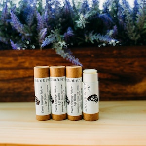 May include: Four lip balm tubes with labels that read "Sweet Orange Tallow Lip Balm" and "Rooted in Her 11:11". The tubes are brown with white labels. A wooden box with purple flowers is in the background.