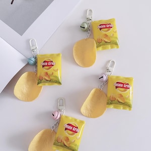 May include: Yellow keychains featuring miniature potato chip bags and a single chip. Each keychain has a silver clasp, a small bell, and the words "Potato Crisp" on the bag. The chips are a light yellow color.
