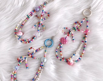 May include: Two colorful beaded phone charms with personalized names, "LESLIE" and "CRYSTAL", and a third charm with the name "KELSEY". The charms feature colorful beads, small bows, and star-shaped charms.