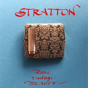 May include: A gold-colored compact with a black and gold lace design. The compact has a small, round, gold-colored container inside. The text "STRATTON" is written in red above the compact. The text "Rare Vintage BEAUTY." is written in red below the compact.