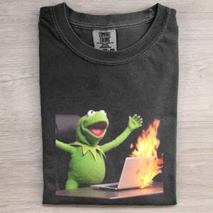 Könnte beinhalten: Schwarzes T-Shirt mit einer Grafik von Kermit dem Frosch mit erhobenen Armen, der an einem Schreibtisch mit einem brennenden Laptop sitzt. Das Shirt hat einen Rundhalsausschnitt und ein Comfort Colors-Etikett.