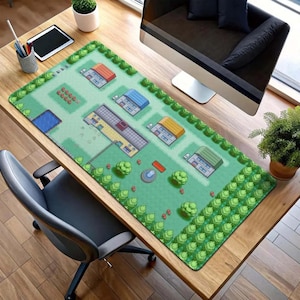 May include: A green rectangular desk mat featuring a pixelated map design with houses, trees, and a pond. The mat is on a wooden desk, with a computer monitor, tablet, and office supplies visible.