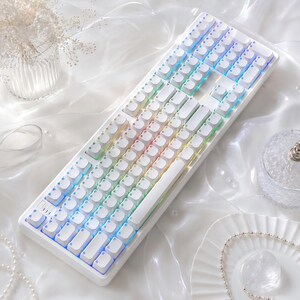 以下が含まれることがあります： 白いキーキャップを備えたフルレイアウトの白いメカニカルキーボードで、キーの間に柔らかな光を放つRGBバックライトが統合されています。キーボードは長方形のフレームと標準的な英数字キーの配置を特徴としています。
