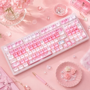 以下が含まれることがあります： 白いフレームとピンクのグラデーションキーキャップを備えたメカニカルキーボード。キーキャップには猫のイラストや肉球のアイコンが描かれています。テンキーとファンクションキーを備え、全体が柔らかなピンク色のバックライトで照らされています。