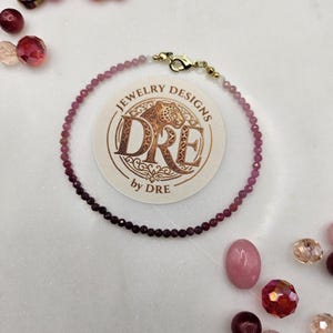 May include: A delicate bracelet with small, round, pink and purple beads. The bracelet is connected with a gold clasp. The image also shows loose beads in shades of pink, purple, and clear, along with a circular logo that reads "JEWELRY DESIGNS by DRE."
