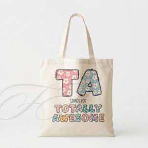 May include: A beige canvas tote bag featuring the text TA STANDS FOR TOTALLY AWESOME. The letters are filled with a colorful floral pattern in shades of pink, blue, yellow, and purple. The design has a dark outline and a stitched appearance.