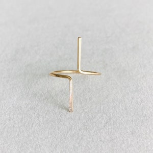 Puede incluir: Un anillo de oro con un diseño simple y minimalista que presenta una forma de cruz formada por dos líneas que se cruzan.