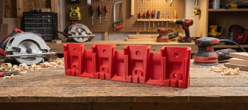 May include: A red power tool battery holder sits on a wooden workbench. The holder has five slots and is surrounded by sawdust and other tools, including a circular saw and a sander. The background includes a pegboard with tools.