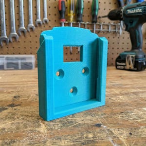 May include: A bright turquoise 3D-printed tool holder with a rectangular opening and three circular holes. The holder is designed to mount on a wall or surface. The background includes tools and a drill.