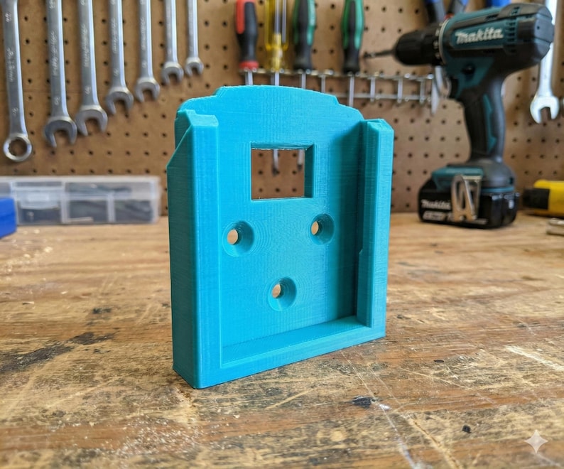 May include: A bright turquoise 3D-printed tool holder with a rectangular opening and three circular holes. The holder is designed to mount on a wall or surface. The background includes tools and a drill.