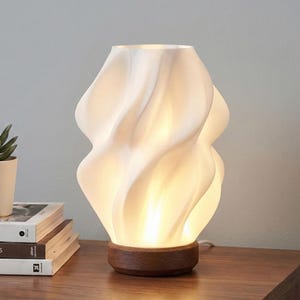 Puede incluir: Una lámpara de mesa blanca y escultural con un diseño orgánico y arremolinado. La lámpara se asienta sobre una base redonda de madera oscura. La lámpara está encendida, emitiendo un brillo cálido. La lámpara está sobre una mesa de madera, junto a una pila de libros.