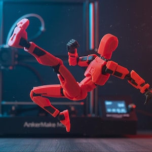 Puede incluir: Una figura articulada roja, con detalles negros en las articulaciones, en plena acción. El fondo borroso muestra una impresora 3D. La escena está iluminada con luces rojas y azules, creando un efecto dinámico y llamativo.