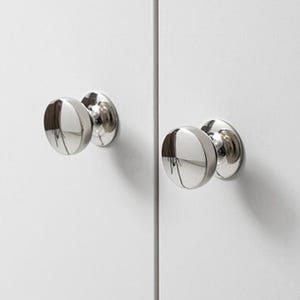 以下が含まれることがあります： 白いキャビネットドアに取り付けられた、2つの磨かれたクロムキャビネットノブ。ノブは、反射面を持つ丸みを帯びた半球状のデザインです。キャビネットのドアはプレーンで白く、それらの間に垂直な縫い目があります。
