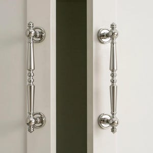 Puede incluir: Dos tiradores de puerta plateados pulidos con un diseño decorativo están montados en una puerta blanca. Los tiradores están orientados verticalmente y tienen un aspecto clásico y elegante. La puerta está ligeramente entreabierta.