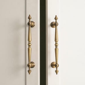 May include: Two brass door pulls with an ornate, elongated design are mounted on a white door. The pulls are vertically oriented and positioned on either side of the door's opening. The hardware adds a touch of elegance to the door.