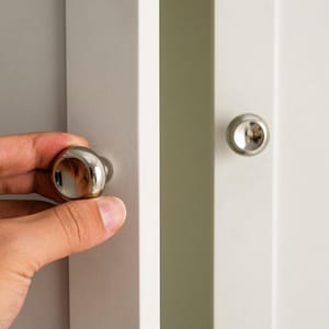 May include: A hand opening a white cabinet door with a shiny, silver knob. The cabinet door is next to a gray cabinet door. A second silver knob is visible on the white door.