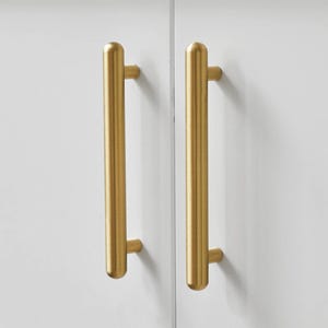Puede incluir: Dos tiradores dorados montados en una puerta de armario blanca. Los tiradores son cilíndricos con extremos redondeados. El armario forma parte de un entorno de cocina o baño, con platos blancos visibles en el fondo.