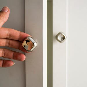 May include: A close-up shot of a white cabinet door with a silver, diamond-shaped knob. The knob reflects the surroundings. Another similar knob is visible on a nearby door. The adjacent door is light grey.