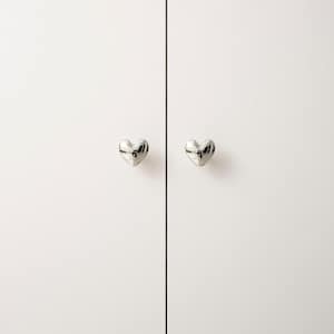 May include: A pair of silver heart-shaped knobs on a white cabinet. The knobs are positioned symmetrically on the cabinet doors, offering a decorative element. The cabinet doors are a plain white colour.