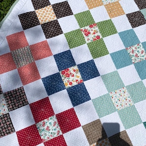 K&ouml;nnte beinhalten: Ein Patchwork-Quilt mit einem Gitter aus wei&szlig;en Quadraten, die sich mit farbig gemusterten Stoffquadraten in Rot-, Blau-, Gr&uuml;n-, Braun- und Gelbt&ouml;nen abwechseln. Der Quilt weist auf der Oberfl&auml;che ein feines, sternf&ouml;rmiges Steppmuster auf.