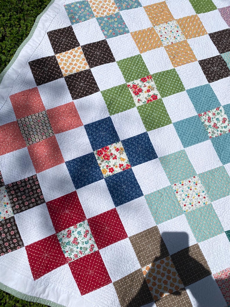 K&ouml;nnte beinhalten: Ein Patchwork-Quilt mit einem Gitter aus wei&szlig;en Quadraten, die sich mit farbig gemusterten Stoffquadraten in Rot-, Blau-, Gr&uuml;n-, Braun- und Gelbt&ouml;nen abwechseln. Der Quilt weist auf der Oberfl&auml;che ein feines, sternf&ouml;rmiges Steppmuster auf.
