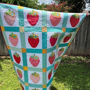 May include: A handmade quilt featuring a grid pattern of white squares, each decorated with a fabric strawberry appliqu&eacute;. The strawberries vary in red, pink, and orange patterns, topped with green leaves and yellow flowers. Teal and yellow fabric strips separate the squares.