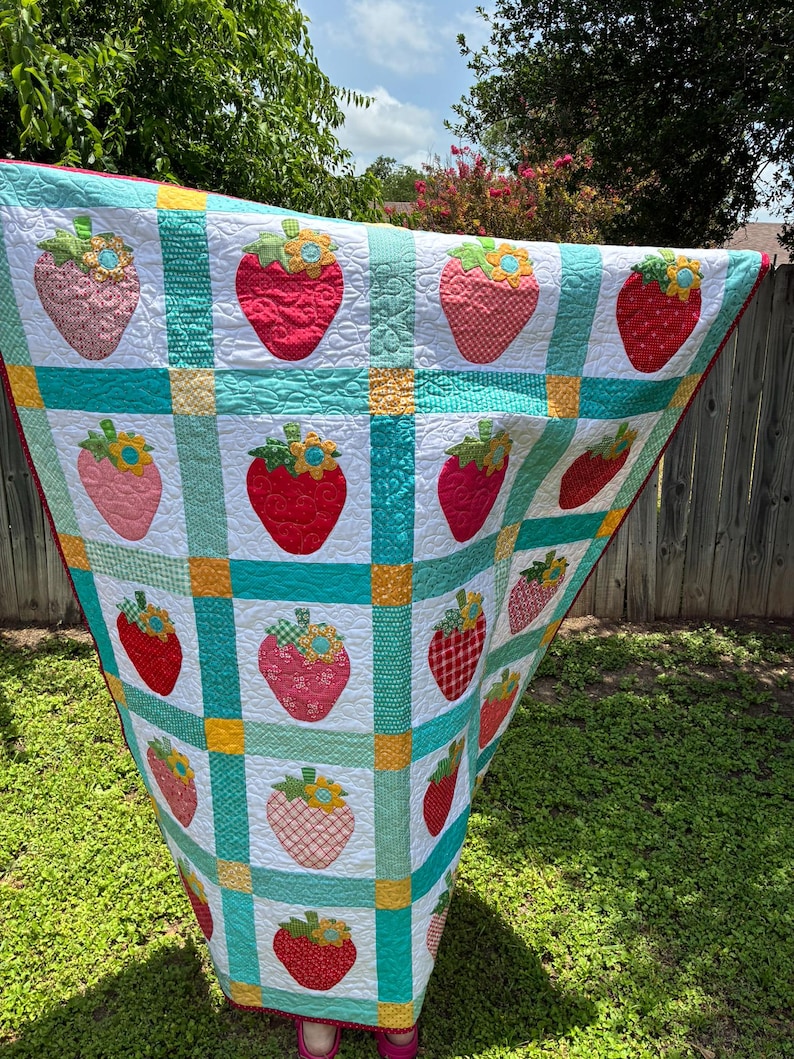 May include: A handmade quilt featuring a grid pattern of white squares, each decorated with a fabric strawberry appliqu&eacute;. The strawberries vary in red, pink, and orange patterns, topped with green leaves and yellow flowers. Teal and yellow fabric strips separate the squares.