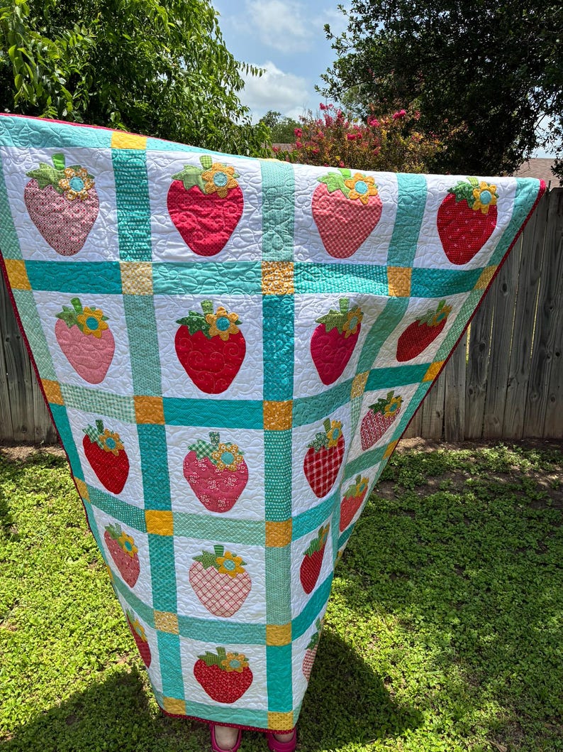 May include: A handmade quilt featuring a grid pattern of white squares, each decorated with a fabric strawberry appliqu&eacute;. The strawberries vary in red, pink, and orange patterns, topped with green leaves and yellow flowers. Teal and yellow fabric strips separate the squares.