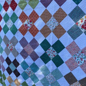 K&ouml;nnte beinhalten: Eine Patchwork-Decke mit einem sich wiederholenden Muster aus wei&szlig;en Quadraten, die mit bunten Stoffquadraten durchsetzt sind. Die farbigen Quadrate zeigen verschiedene Blumenmuster, Punkte und einfarbige Texturen in Rot, Gr&uuml;n, Blau und Braun.