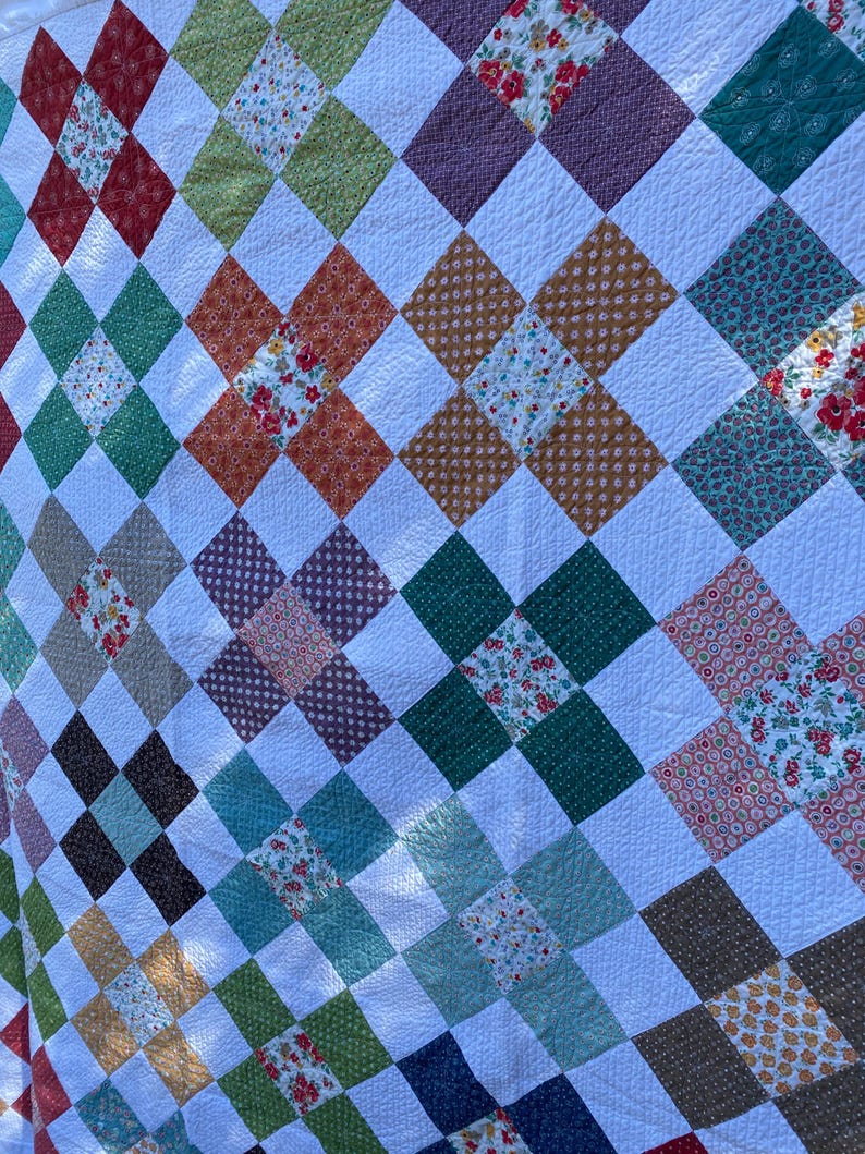 K&ouml;nnte beinhalten: Eine Patchwork-Decke mit einem sich wiederholenden Muster aus wei&szlig;en Quadraten, die mit bunten Stoffquadraten durchsetzt sind. Die farbigen Quadrate zeigen verschiedene Blumenmuster, Punkte und einfarbige Texturen in Rot, Gr&uuml;n, Blau und Braun.