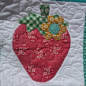May include: A quilted fabric square featuring a central red strawberry applique with a white floral pattern. The strawberry has a green gingham leaf, a small green floral stem, and a yellow flower with a blue center. The background is white fabric.