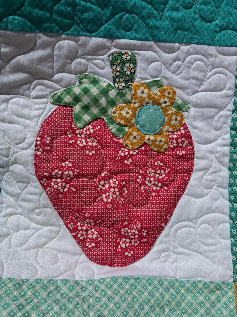 May include: A quilted fabric square featuring a central red strawberry applique with a white floral pattern. The strawberry has a green gingham leaf, a small green floral stem, and a yellow flower with a blue center. The background is white fabric.