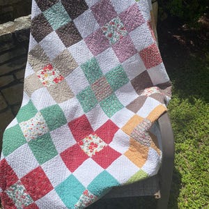 K&ouml;nnte beinhalten: Eine Patchwork-Decke mit einem sich wiederholenden Blockmuster aus wei&szlig;en Quadraten, durchsetzt mit Quadraten in verschiedenen Farben wie Rot, Gr&uuml;n, Braun, Gelb und Rosa. Der Stoff zeigt diverse Blumen- und geometrische Drucke mit sichtbarer Steppung.