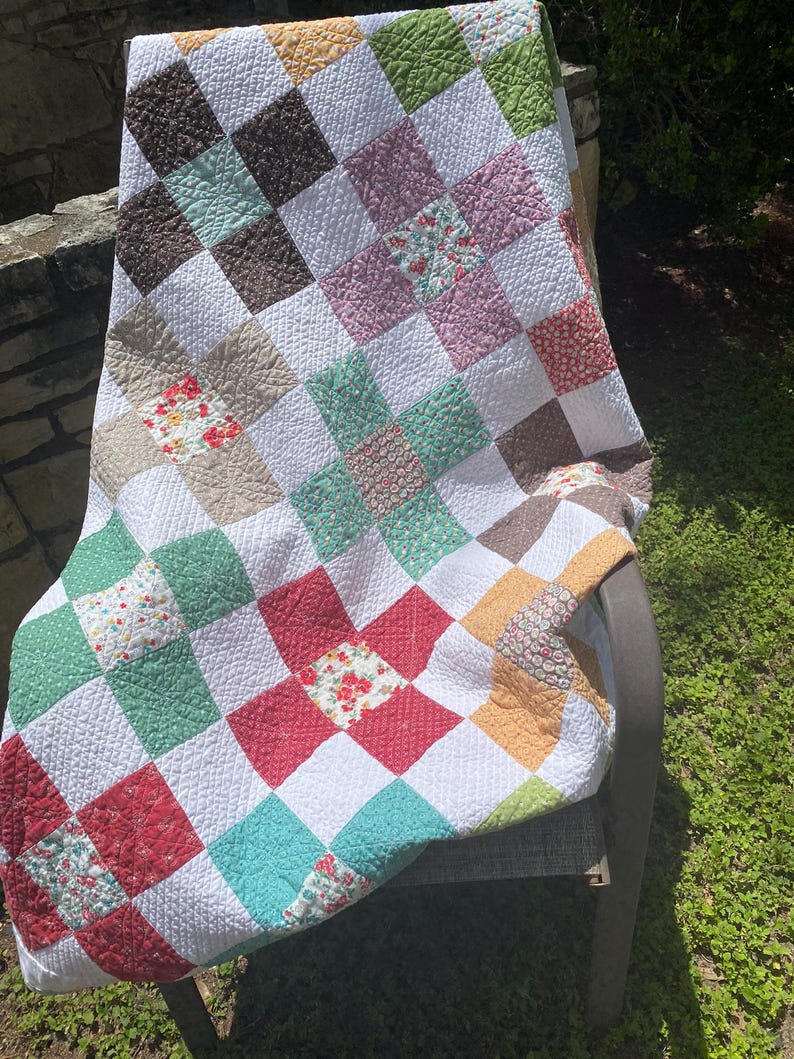 K&ouml;nnte beinhalten: Eine Patchwork-Decke mit einem sich wiederholenden Blockmuster aus wei&szlig;en Quadraten, durchsetzt mit Quadraten in verschiedenen Farben wie Rot, Gr&uuml;n, Braun, Gelb und Rosa. Der Stoff zeigt diverse Blumen- und geometrische Drucke mit sichtbarer Steppung.