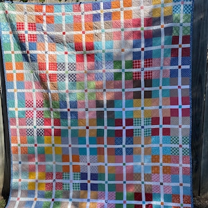 Może przedstawiać: Kolorowa patchworkowa kołdra z wzorem kratki. Kołdra składa się z kwadratów w różnych kolorach, w tym czerwonym, niebieskim, żółtym i zielonym, oddzielonych białymi paskami. Kołdra ma niebieską obwódkę.