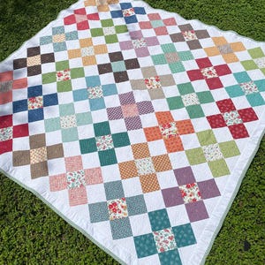 K&ouml;nnte beinhalten: Eine quadratische Patchwork-Decke mit einem sich wiederholenden Kreuzmuster aus verschiedenen bunten Stoffquadraten. Das Design umfasst Blumen-, Punkte- und Unimuster in Rot-, Blau-, Gr&uuml;n-, Orange- und Violettt&ouml;nen auf wei&szlig;em Hintergrund mit einem hellgr&uuml;nen Rand.